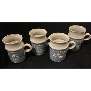 May include: Four ceramic mugs with a two-tone design. The top portion is a light beige, while the bottom is a muted blue with vertical lines and white floral accents. Each mug has a curved handle and a slightly flared rim.