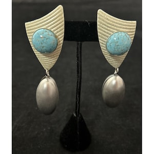 May include: A pair of unique earrings. Each earring features a light beige, shield-shaped top with a textured design and a turquoise-colored stone. A silver-colored, oval-shaped charm dangles from the bottom of each earring. The earrings are displayed on a black stand.