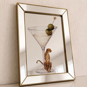 Può includere: Uno specchio rettangolare con cornice dorata che presenta un'illustrazione stampata di un bicchiere da martini contenente due olive su uno stuzzicadenti con una stella in cima, e un ghepardo seduto alla base del gambo del bicchiere.