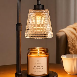 May include: A black metal candle warmer lamp with a textured clear glass shade. Beneath the lamp sits a glass jar candle labeled Solar Good Coconut + Vanilla, Net Wt. 10 oz | 283 g. The lamp emits a warm glow, illuminating the candle jar below.