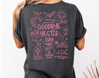 May include: Dark gray t-shirt with a pink graphic design. The design includes the words "Goodbye Yesterday Day" and various religious symbols and phrases, such as a cross, a road, and "I have decided to follow Jesus."