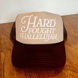 May include: A brown and beige trucker hat with a white embroidered design that says "Hard Fought Hallelujah".