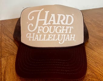 May include: A brown and beige trucker hat with a white embroidered design that says "Hard Fought Hallelujah".