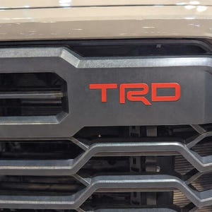 May include: Close-up of a vehicle's front grille. The dark grey grille features the red letters "TRD" prominently displayed. The grille has a geometric design with horizontal and angled bars, and a tan-coloured vehicle body is visible above.