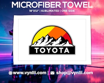 May include: White microfiber towel, 18" x 12", with a mountain and sun logo above the Toyota text. The logo features a black mountain range against a yellow and orange gradient. The towel is sublimated on one side.