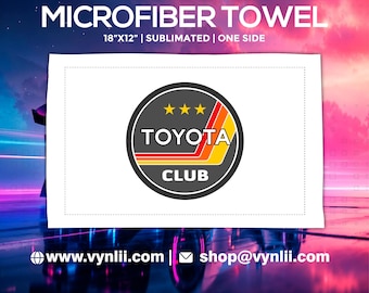 May include: Microfiber towel, 18" x 12", with a sublimated Toyota Club logo. The logo is a circular design with the word "TOYOTA" and "CLUB" in white, and a red, orange, and yellow stripe design. The towel is white.