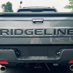 May include: The rear view of a gray pickup truck tailgate featuring the word RIDGELINE embossed in large, dark, block letters. The vehicle includes a black bumper with integrated step pads, two circular exhaust pipes, and a central handle.