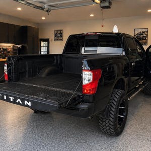 May include: A black pickup truck with the tailgate down, displaying the word "TITAN" in white. The truck has large, black wheels and is parked in a garage. The door is open, and the truck bed is visible.