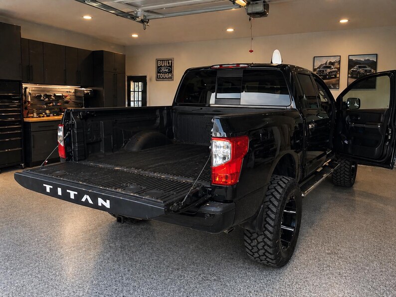 May include: A black pickup truck with the tailgate down, displaying the word "TITAN" in white. The truck has large, black wheels and is parked in a garage. The door is open, and the truck bed is visible.