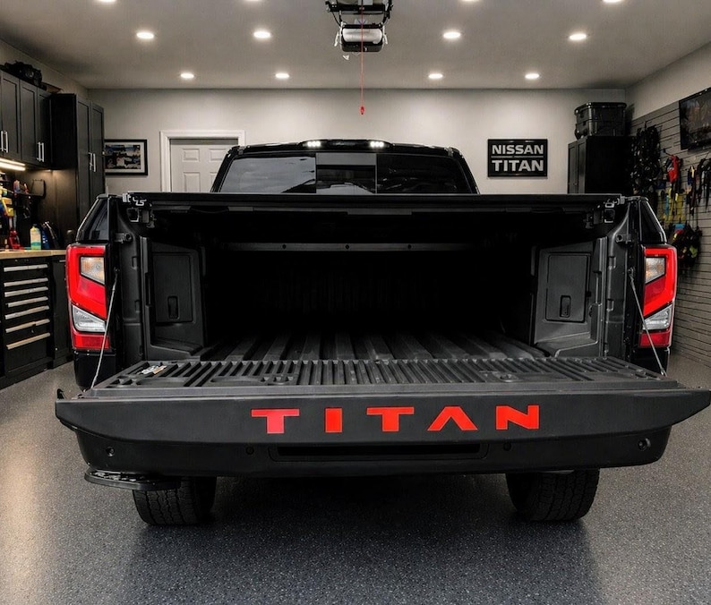 May include: Black Nissan Titan pickup truck with the tailgate open, displaying the red "TITAN" lettering. The truck bed is visible, and the taillights are illuminated. The truck is parked inside a garage.