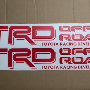 May include: Two red TRD Off Road Toyota Racing Development decals on a white background. The decals are on a cardboard surface.