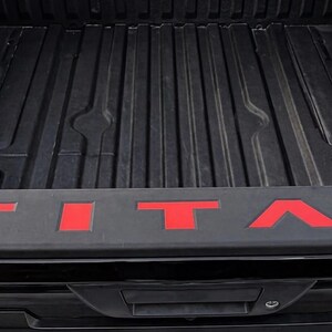 May include: Black truck bed with the red word "TITAN" across the tailgate. The bed has a ribbed design. The tailgate has a handle and a lock.