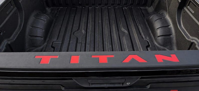 May include: Black truck bed with the red word "TITAN" across the tailgate. The bed has a ribbed design. The tailgate has a handle and a lock.