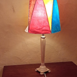 May include: A colourful table lamp with a geometric lampshade. The shade is made up of red, yellow, blue, and brown panels. The lamp has a cream-coloured base and stem, and sits on a dark wooden surface.