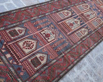 May include: A rectangular, hand-woven rug with a detailed geometric pattern. The rug features a central design in shades of blue, red, and beige, framed by a border of intricate patterns. Fringes are visible at the ends, adding to its traditional aesthetic.