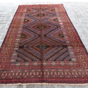 Pode incluir: Um tapete retangular tecido à mão com um padrão geométrico. O tapete apresenta um design central em forma de diamante em tons de castanho, azul e vermelho. A borda é de um vermelho mais escuro com motivos geométricos repetidos.