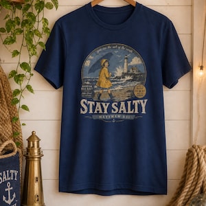 May include: A navy blue short-sleeve t-shirt featuring a graphic print of a person in a yellow raincoat walking on a beach near a lighthouse. The text reads "STAY SALTY" and "MATTHEW 5:13" along with "PREMIUM SEA SALT" and "you are the salt of the earth."