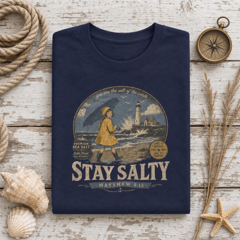 May include: A navy blue t-shirt featuring a graphic of a child in a yellow raincoat holding an umbrella near a lighthouse. The text reads: "you are the salt of the earth", "PREMIUM SEA SALT", "STAY SALTY", and "MATTHEW 5.13" in a vintage style.