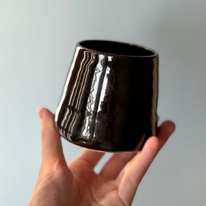May include: A black ceramic cup with a glossy finish and a gently widening form. The cup is held in a hand against a pale blue backdrop. The surface reflects light, emphasizing its smooth texture and dark hue.