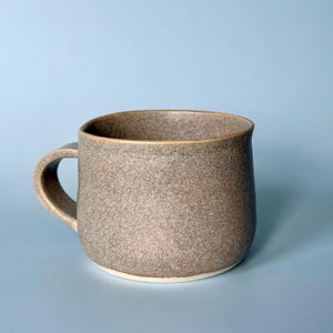 May include: A speckled, grey ceramic mug with a rounded body and a curved handle. The mug has a light-coloured rim and base, set against a light blue background. This handmade mug is suitable for coffee, tea, or other beverages.