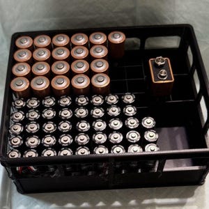 May include: A black plastic battery organizer filled with various batteries. The organizer holds numerous AA and AAA batteries, along with a 9-volt battery. The batteries are arranged neatly within the compartments of the organizer.