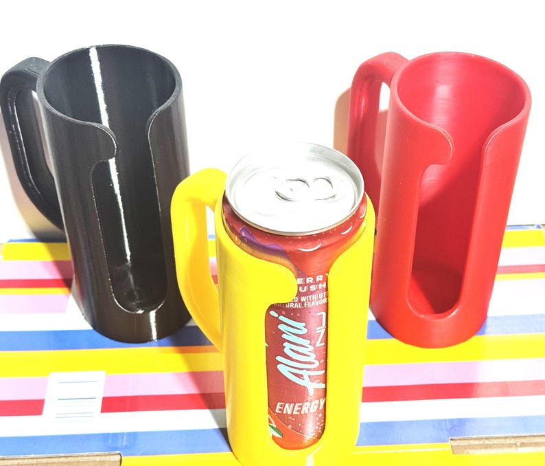 May include: Three 3D-printed can holders in black, yellow, and red. The yellow holder contains a red and silver can with the text "Alani Energy". Each holder has a handle for easy carrying. The holders are designed to keep cans secure.