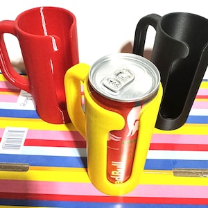 Puede incluir: Tres soportes para latas impresos en 3D en rojo, amarillo y negro. El soporte amarillo contiene una lata de Red Bull. Cada soporte tiene un asa y un dise&ntilde;o curvo para sujetar de forma segura una lata. Los soportes est&aacute;n sobre una superficie a rayas colorida.