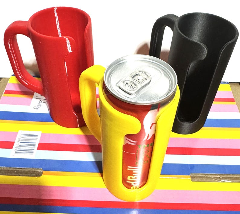 Puede incluir: Tres soportes para latas impresos en 3D en rojo, amarillo y negro. El soporte amarillo contiene una lata de Red Bull. Cada soporte tiene un asa y un dise&ntilde;o curvo para sujetar de forma segura una lata. Los soportes est&aacute;n sobre una superficie a rayas colorida.