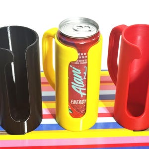 May include: Three 3D-printed can holders in black, yellow, and red. The yellow holder contains a can of Alani Energy drink. Each holder has a handle and a cylindrical design to securely hold a standard-sized beverage can.