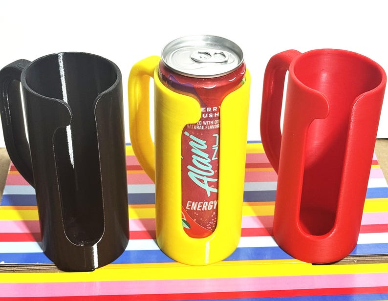 May include: Three 3D-printed can holders in black, yellow, and red. The yellow holder contains a can of Alani Energy drink. Each holder has a handle and a cylindrical design to securely hold a standard-sized beverage can.