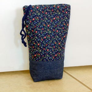 May include: A tall, fabric drawstring bag with a floral pattern on a navy blue background. The bottom section is a solid dark denim. A navy blue drawstring is attached to the top.