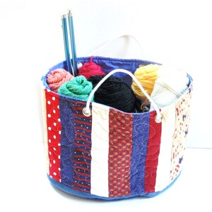 May include: A round, patchwork fabric basket filled with colorful yarn balls and two blue knitting needles. The basket features red, white, and blue patterned fabric strips, with a white handle. The interior is blue.