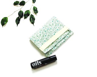 Pode incluir: Uma bolsa de tecido retangular com um padrão floral pequeno em verde e branco e um fecho de elástico na cor creme, acompanhada por um frasco cilíndrico preto com o texto branco "oils" impresso em uma fonte em negrito.