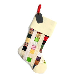 Pode incluir: Uma meia de Natal artesanal com um design de patchwork. A meia apresenta um punho e uma ponta de cor creme, com um corpo composto por v&aacute;rios quadrados de tecido em diferentes cores e padr&otilde;es. Uma fita vermelha e uma etiqueta preta est&atilde;o presas.