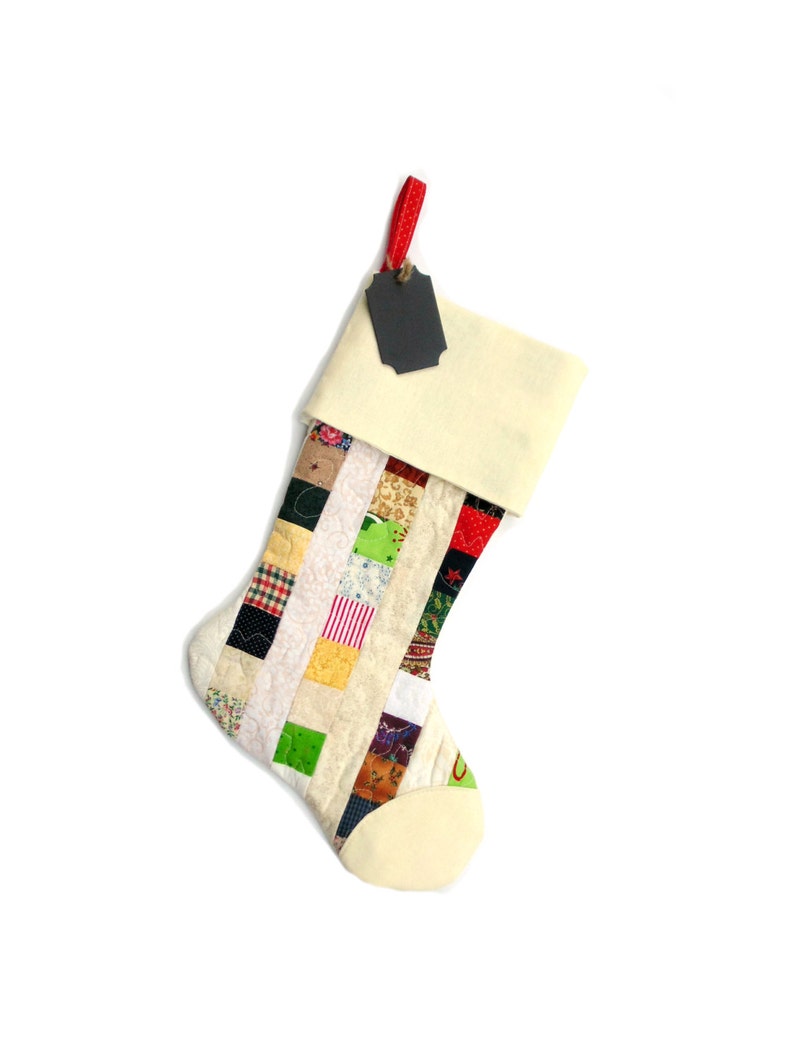 Pode incluir: Uma meia de Natal artesanal com um design de patchwork. A meia apresenta um punho e uma ponta de cor creme, com um corpo composto por v&aacute;rios quadrados de tecido em diferentes cores e padr&otilde;es. Uma fita vermelha e uma etiqueta preta est&atilde;o presas.