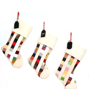 Pode incluir: Tr&ecirc;s meias de Natal com um design de patchwork. Cada meia tem um punho e uma ponta creme, com um corpo de patchwork colorido. Etiquetas pretas penduradas em barbante vermelho.