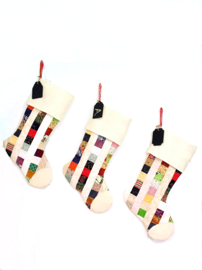 Pode incluir: Tr&ecirc;s meias de Natal com um design de patchwork. Cada meia tem um punho e uma ponta creme, com um corpo de patchwork colorido. Etiquetas pretas penduradas em barbante vermelho.
