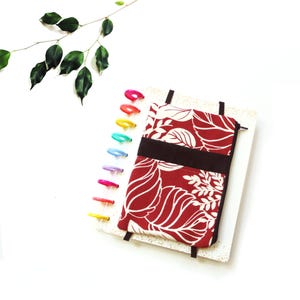 May include: A red and white floral patterned notebook cover with a dark brown band and black straps. The notebook is open, revealing white pages. Colorful plastic disc-bound rings are visible on the left side.