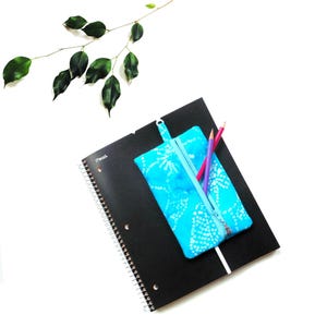 May include: A black spiral-bound notebook with a blue patterned pencil case and two colored pencils. The pencil case has a zipper and a loop. A sprig of green leaves is in the upper left corner. The pencil case is filled with two colored pencils.