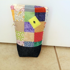 May include: A patchwork fabric sleeve, likely for a bottle or container, with a denim base. The sleeve is composed of various fabric squares in colors like yellow, green, purple, and red. A yellow square with a black design is visible.