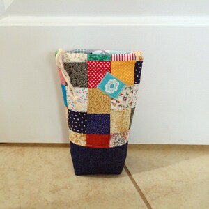 May include: A patchwork fabric doorstop with a denim base. The body of the doorstop is made of various colorful fabric squares, including polka dots, floral prints, and solid colors. A drawstring is attached to the top.