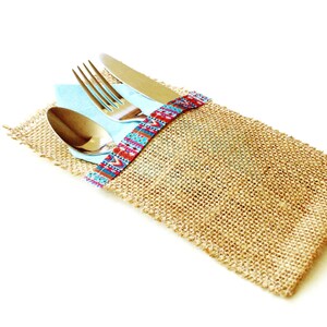 May include: A burlap napkin holder with a blue and red patterned ribbon. The holder contains a silver fork, knife, and spoon.