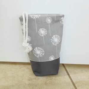 May include: A gray fabric bag with a white dandelion pattern and a dark gray bottom. The bag has a white drawstring closure at the top. The bag is standing upright on a tiled surface.
