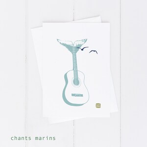 May include: A white greeting card with a teal guitar illustration topped with a whale tail and two flying birds. The words "chants marins" are printed in green at the bottom. The card is placed on a white surface.