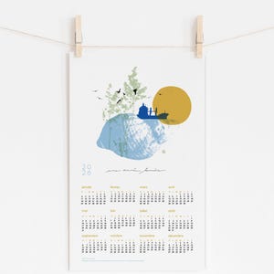 May include: A 2026 calendar print, featuring an abstract landscape with a blue and yellow color palette. The design includes a silhouette of a building, trees, birds, and a sun. The calendar is held by wooden clothespins.