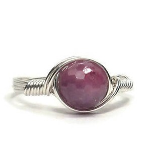 May include: A silver wire-wrapped ring featuring a round, polished, deep pink gemstone. The gemstone is the focal point, set in a delicate, handcrafted design. The ring is displayed on a white background, highlighting the jewelry's details.