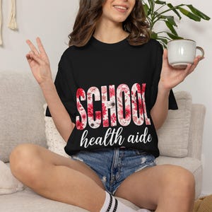 Puede incluir: Camiseta negra con las palabras "SCHOOL" y "health aide" en un diseño floral. La camiseta se usa con pantalones cortos de mezclilla. Una mujer sostiene una taza y hace el signo de la paz.