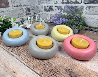 May include: Five smooth, rounded concrete candle holders in pastel blue, light grey, pale yellow, and soft pink. Each holder features a circular centre cutout containing a small, round yellow tea light candle with a visible cotton wick.