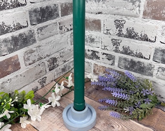 May include: A tall, smooth, dark green taper candle stands upright in a round, light grey concrete candle holder. The candle has a pointed top with a visible white wick. The base of the holder is circular with a raised centre to secure the candle.