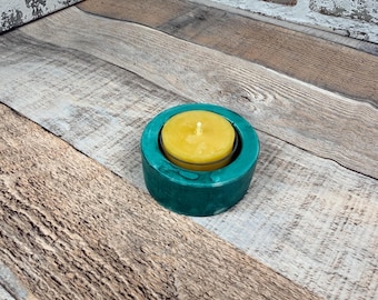 May include: A circular teal candle holder with a smooth, polished finish containing a single round yellow wax tea light candle with a visible white wick centred in the middle of the holder's recessed circular opening.